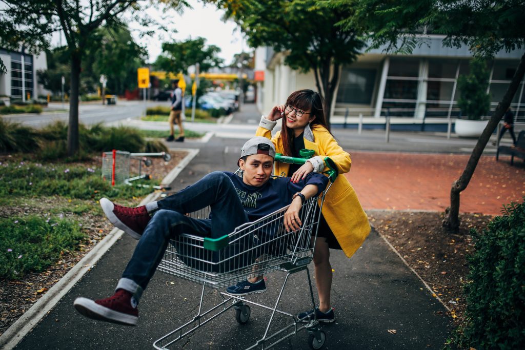 A woman leans onto a shopping cart, while a man poses inside on a city sidewalk